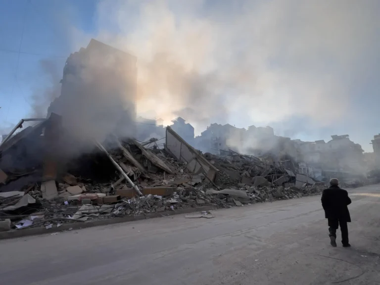 A person walks along a dusty road past a massive pile of building rubble and rising smoke under a hazy sky.
